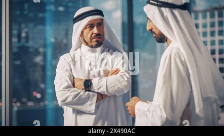 Zwei erfolgreiche muslimische Geschäftsleute in weißem Traditional Outfit, die im Büro sitzen, verhandeln und über finanzielle Chancen sprechen. Saudi, Emirat, Arabisches Geschäftsmann-Konzept. Stockfoto
