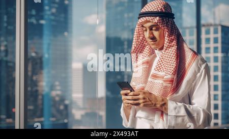 Junger muslimischer Geschäftsmann im traditionellen weißen Kandura in seinem modernen Büro, mit Smartphone neben Fenster mit Wolkenkratzern. Erfolgreiches Konzept Für Saudi-Arabien, Emirate, Arabische Geschäftsleute. Stockfoto