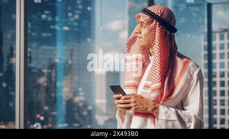 Junger muslimischer Geschäftsmann im traditionellen weißen Kandura in seinem modernen Büro, mit Smartphone neben Fenster mit Wolkenkratzern. Erfolgreiches Konzept Für Saudi-Arabien, Emirate, Arabische Geschäftsleute. Stockfoto