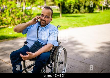 Porträt des glücklichen Mannes im Rollstuhl. Er genießt einen sonnigen Tag im Stadtpark und spricht am Telefon. Stockfoto