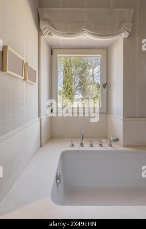 Eine Badewanne aus weißem Porzellan mit Marmorumrandung mit Armaturen im Vintage-Stil und einem weißen Aluminiumfenster mit Blick auf den Wald Stockfoto