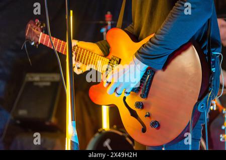 Ein Musiker spielt auf der Bühne eine orangene, halbhohlige elektrische Gitarre mit hellgelben Lichtern im Hintergrund. Der Gitarrist trägt eine Jacke Stockfoto