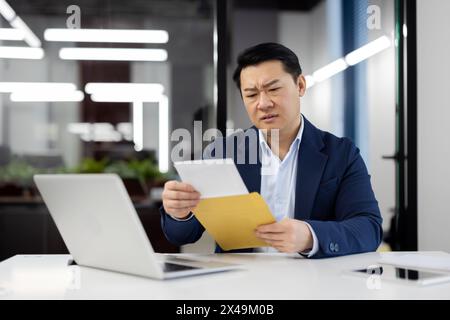 Gestörter asiatischer Mann im Business-Anzug, der den Brief aus dem Umschlag herausnimmt, während er mit pc im Schrank sitzt. Gestresster Unternehmer, der postalische Benachrichtigungen erhält Stockfoto