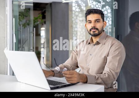 Ernsthafter islamischer Geschäftsmann, der während des Arbeitstages am Arbeitsplatz mit Computer die Brille abzieht. Konzentrierter Startup-Läufer, der harte Entscheidungen über die Wachstumsstrategie des Unternehmens trifft. Stockfoto