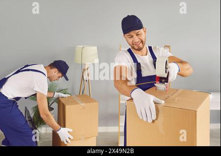 Zwei professionelle junge männliche Mover sichern Kartons mit Klebeband in einem neuen Zuhause. Stockfoto