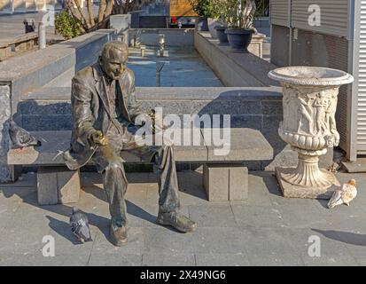 Craiova, Rumänien - 16. März 2024: Bronzestatue des Menschen, der Tauben füttert, sitzt auf einer Bank im Stadtzentrum am sonnigen Frühlingstag. Stockfoto