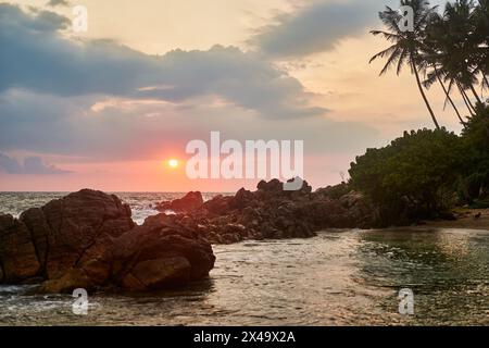 Sonnenuntergang über tropischem Strand mit Felsen, ruhiges Meer. Palmen Silhouette gegen Abendhimmel. Goldene Sonne am Horizont reflektiert auf dem Wasser. Friedlich Stockfoto