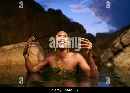 Lächeln des lgbtq-Modells im Wasser im natürlichen Pool bei Nacht. Nicht binäre Person zeigt Schmuck - Ringe mit Edelsteinen an den Fingern, Messingring Stockfoto