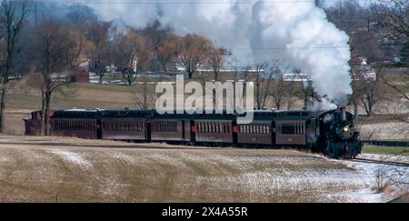 Eine Dampfeisenbahn fährt eine verschneite Strecke entlang. Der Zug ist schwarz-weiß und von Bäumen umgeben. Der Zug ist der Hauptfokus des Bildes, und Stockfoto