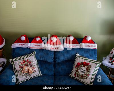 Eine Gruppe von fünf roten weihnachtsmännern sitzt auf einer blauen Couch. Die Hüte sind mit Namen personalisiert und in einer Reihe angeordnet. Die Couch ist dekoriert Stockfoto