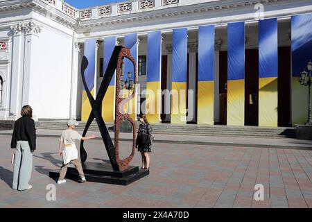 Odessa, Ukraine. Mai 2024. Die Menschen betrachten die skulpturale Komposition „EPIPHANY“ auf dem Dumskaja-Platz. Die skulpturale Komposition „EPIPHANY“ (Stahl und Kupfer) von Mikhail Reva entstand 2024 und stellt einen Rahmen für Gläser dar. Einige der Kugeln sind in Form von Blumen dargestellt, denn wenn sie im menschlichen Körper platzen, verwandeln sie sich in Elemente ähnlicher Form. Es gibt eine andere Bedeutung, die dem Werk innewohnt: Dies sind die Buchstaben „XB“, die von der Skulptur gebildet werden. Nach Ansicht des Autors handelt es sich dabei nicht um einen Zufall, sondern um eine Bezugnahme auf den Christen „Christus ist auferstanden“ als Symbol Stockfoto