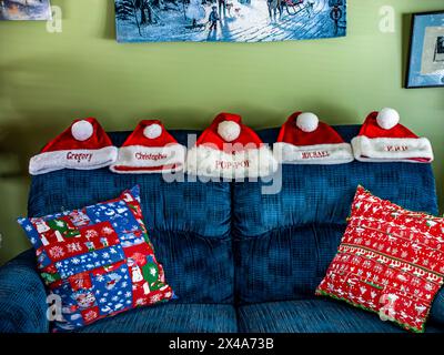 Eine Gruppe von fünf roten weihnachtsmännern sitzt auf einer blauen Couch. Die Hüte sind mit Namen personalisiert und in einer Reihe angeordnet. Die Couch ist mit einem rot-weißen Kissen und einem blauen Kissen dekoriert Stockfoto