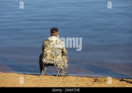 Fischer, der am Wasser sitzt, mit einer Angelrute, Rückansicht. Mann, der im Frühling an der Flussküste angeln kann Stockfoto