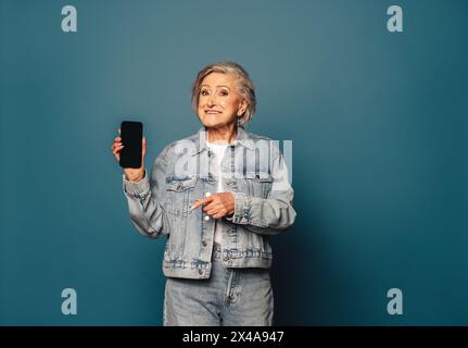 Porträt einer fröhlichen, Reifen Frau in Denim-Kleidung, die in einem Studio steht und ein Smartphone mit der Hand hält. Sie lächelt, während sie den Bildschirm zeigt Stockfoto