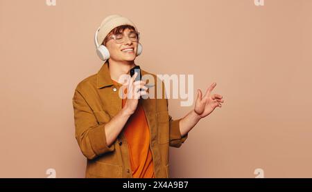 Junger Mann in lässigem Outfit und Jeansjacke steht vor pfirsichfarbenem Hintergrund im Studio. Er singt glücklich zu einem Lied, trägt Brille und Kopfhörer Stockfoto