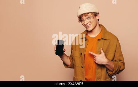 Junger Mann in lässiger Kleidung und Jeansjacke hält ein Handy mit einer mobilen App. Er zeigt auf den Touchscreen und empfiehlt ihn fröhlich. Das vibr Stockfoto
