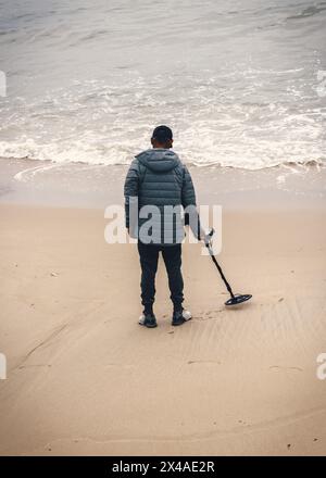 Ein einsamer, nicht identifizierbarer Mann steht an einem Sandstrand und hält einen Metalldetektor, während er die Küste nach versteckten Schätzen durchsucht Stockfoto