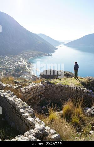 Ein Mann steht auf der Spitze in der Nähe der antiken Ruinen, vor dem Hintergrund eines malerischen Ausblicks auf das Meer und die Berge (Risan, Montenegro) Stockfoto
