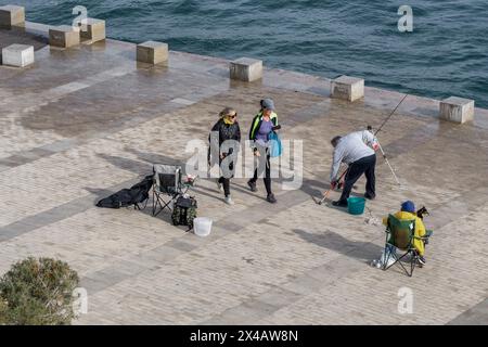 Zwei Frauen gehen, eine sitzend und ein Mann legt die Angelrute im Hafen an der Promenade des Weihnachtsleuchtturms in Cartagena. Stockfoto