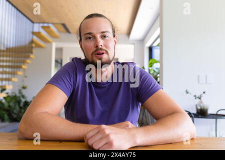 Kaukasischer junger Mann mit Bart, der zu Hause am Holztisch sitzt, während eines Videoanrufs Stockfoto