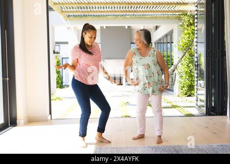 Asiatische Großmutter und birassische Teenager-Enkelin tanzen zu Hause zusammen. Beide barfuß, tragen legere Kleidung, teilen freudige Momente, unaltere Stockfoto