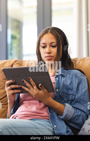 Zu Hause ein birassisches Teenager-Mädchen, das Kopfhörer trägt und ein Tablet hält. Sie hat hellbraune Haut und langes dunkles Haar, kleidet sich lässig und genießt Musik Stockfoto