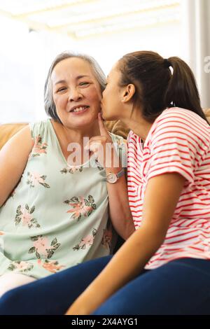 Birassische Teenager-Enkelin, die asiatische Oma zu Hause auf die Wange küsst. Beide tragen legere Kleidung, teilen Zuneigung, sitzen in warmen Momenten nah, u Stockfoto