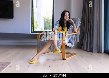 Junge birassische Frau, die sich zu Hause in einem modernen Stuhl entspannt. Sie hat lange dunkle Haare, trägt bunte Kleidung und gelbe Sandalen, unverändert Stockfoto