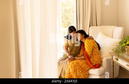 Indische Mutter tröstet Teenager-Tochter auf Sofa, beide in traditioneller Kleidung, Kopierraum. Mit Goldschmuck, Teenager-Tochter mit Teddybär Stockfoto