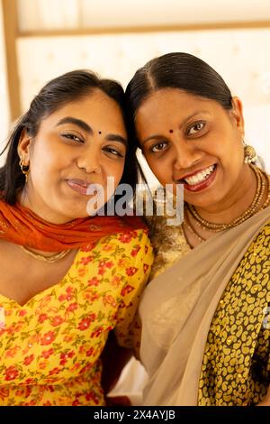 Indische Mutter und Tochter im Teenageralter, die sich zu Hause umarmen und beide lächeln vor der Kamera. Mutter mit dunklem Haar und Bindi, mit Goldschmuck, Teenager-Tochter Stockfoto