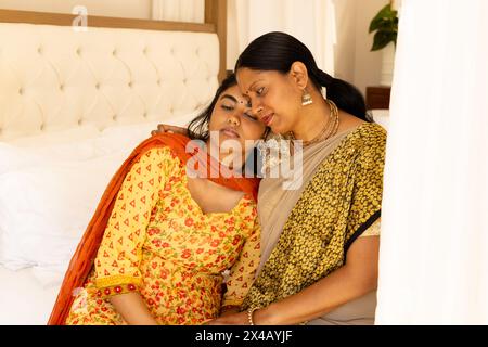 Indische Mutter und Tochter im Teenageralter, die sich im Bett umarmen und traditionelle Kleidung tragen. Beide zeigen Zuneigung, mit langen und mittellangen schwarzen Haaren, unalt Stockfoto