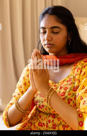 Eine junge Indianerin, betend mit geschlossenen Augen zu Hause, in gelbem Kleid. Orangefarbener Schal, langes schwarzes Haar und goldene Ohrringe, die Ruhe und Ruhe zum Ausdruck bringen Stockfoto