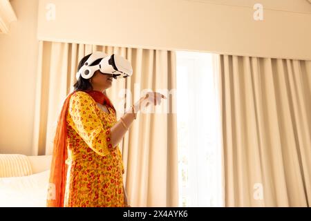Indische junge Frau, die zu Hause ein Virtual-Reality-Headset trägt, im Schlafzimmer steht. Gelbes Kleid mit roten Mustern, mit dem Sie den virtuellen Raum erkunden Stockfoto