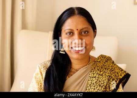 Eine Indianerin mittleren Alters in traditioneller Kleidung, die zu Hause lächelt. Sie hält ihr Smartphone, hat dunkle Haare und braune Augen. Sie genießt den Moment, unverändert Stockfoto