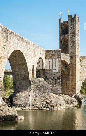 Die Brücke des mittelalterlichen Dorfes Besalú mit einem Turm im Hintergrund. Die Brücke besteht aus Stein und hat einen Steinbogen. Das Wasser ist ruhig und es ist ruhig Stockfoto