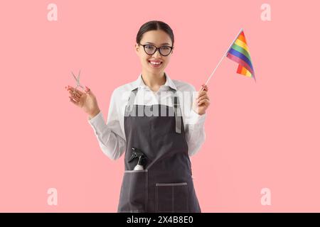 Weiblicher asiatischer Friseur mit Schere und LGBT-Flagge auf rosa Hintergrund Stockfoto