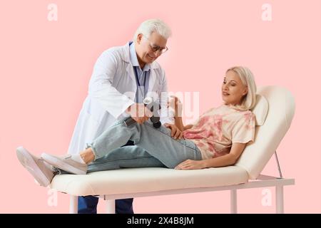 Männlicher Physiotherapeut massiert das Bein der Reifen Frau mit perkussivem Massagegerät auf der Couch vor rosa Hintergrund Stockfoto