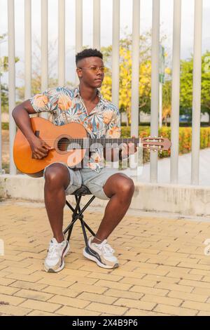 Ein junger afrikanischer Mann spielt seine Gitarre in einem urbanen Park, gekleidet mit einem Hemd mit tropischem Muster und einer stilvollen Sonnenbrille. Stockfoto