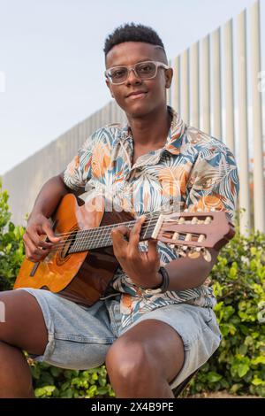 Ein fröhlicher junger afrikanischer Mann in Brille und farbenfrohem Hemd spielt Gitarre, lächelt in die Kamera, vor einem urbanen Hintergrund. Stockfoto