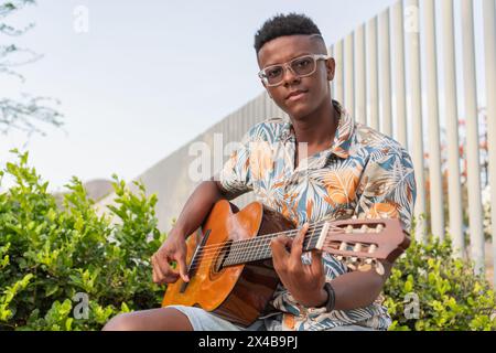 Selbstbewusster junger afrikanischer Mann mit Brille und Blumenhemd, der draußen Akustikgitarre spielt, umgeben von grünem Laub. Stockfoto