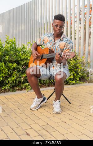 Ein junger afrikanischer Mann in Sonnenbrille und einem lebhaften tropischen Hemd spielt eine Akustikgitarre, während er draußen auf einem Hocker sitzt. Stockfoto