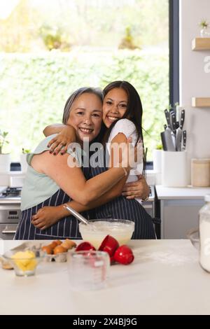 Asiatische Großmutter und birassische Teenager-Enkelin umarmen sich zu Hause in der Küche. Beide lächeln, Senioren mit kurzen grauen Haaren, die Jungen mit Stockfoto