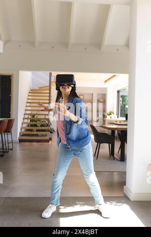 Eine birassische junge Frau, die zu Hause ein VR-Headset trägt und spielt. Mit langen dunklen Haaren, lässigem blauem Hemd und Jeans, eingetaucht in ein virtuelles Spiel Stockfoto