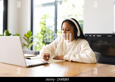 Eine birassische junge Frau, die Kopfhörer trägt und zu Hause Notizen schreibt. Mit langen dunklen Haaren und hellbrauner Haut, sieht sie ihren Laptop, gekleidet Casua Stockfoto