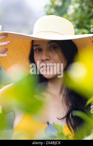 Birassische junge Frau mit gelbem Badeanzug und breiter Krempe draußen. Sie hält ihren Hut und zeigt ein sanftes Lächeln mit dunklen Haaren gegen ein gelbes B Stockfoto