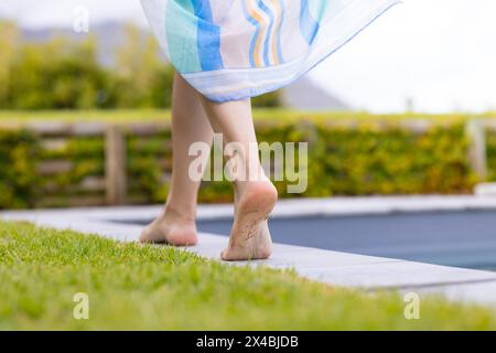 Birassische junge Frau, die ein Handtuch hält, draußen am Pool spaziert. Sie hat hellbraune Haut und dunkles Haar, genießt barfuß einen Spaziergang, unverfälscht Stockfoto