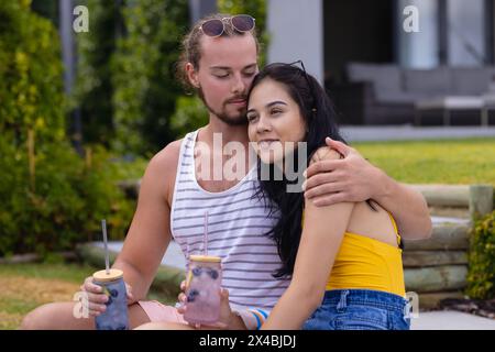 Kaukasier männlich und birassisch weiblich genießen Drinks im Freien. Er trägt Bart und braunes Haar, hält ein Glas, sie mit langen schwarzen Haaren in Gelb Stockfoto