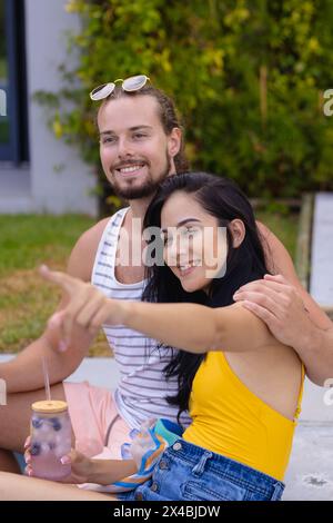 Diverse Paare genießen Drinks draußen am Pool und zeigen auf etwas. Sie hat dunkle Haare, er hat Bart und hellbraune Haare, sowohl lachend als auch holdin Stockfoto