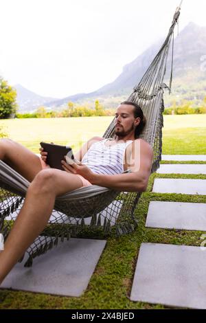 Ein junger kaukasischer Mann, der sich draußen in der Hängematte entspannt, eine Tablette hält, einen Kopierraum. Mit dunklen Haaren und Bart, mit einem Tanktop und Shorts, genießen Stockfoto