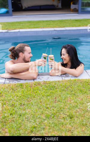 Diverse Paare genießen Drinks draußen am Pool und lachen zusammen. Sie hat dunkle Haare, und er hat lange braune Haare und Bart Stockfoto